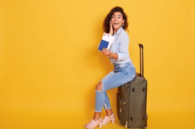 Pasaportu ve bileti olan Avrupalı bir kadın bavulun üzerinde mutlu ve heyecanlı görünüyor, ağzı açık ve avuç içi ile yanağına dokunuyor, kızın yıllık tatili var.