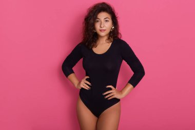 Kalçasında elleri olan esmer bir kadın, dalgalı saçları var, pembe arka planda izole edilmiş poz veriyor, siyah bir elbise giyen çekici bir kız, doğrudan kameraya bakıyor, kendinden emin görünüyor..