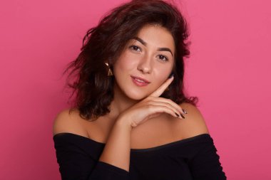 Çıplak omuzlu, şık siyah gömlekli, pembe duvarda poz veren, yanağında parmağını tutan, dalgalı saçlı, doğrudan kameraya bakan bir kız..