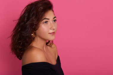 Güzel, koyu renk saçlı, kendine güvenen genç bir bayan, şık bir dergiye poz veriyor, düşünceli bir şekilde kenara bakıyor, çıplak omuzlu, pembe arka planda poz veren şık bir gömlek giyiyor..