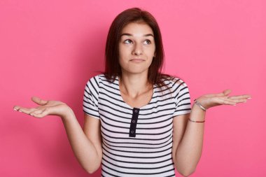 Güvensiz, şaşkın, koyu renk saçlı kadın, kuşkuyla el ele tutuşan, sıradan çizgili tişört giyen, pembe arka plan üzerinde izole edilmiş poz veren, yana bakan.