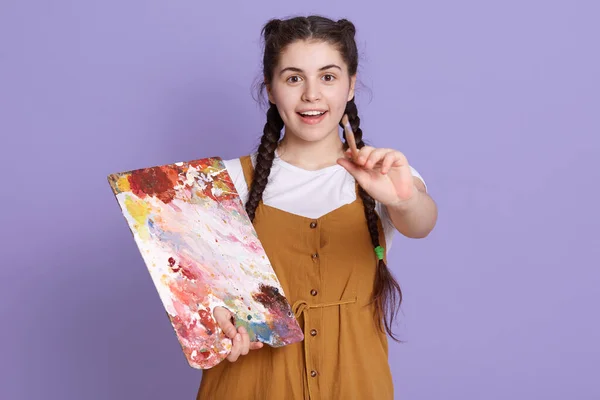 Female artist at work posing isolated lilac wall with excited expression, holding colors palette and paintbrush in hands, girl with pigtails looks smiling at camera.