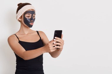 Duştan sonra siyah maskeli genç mutlu bir kadının portresi beyaz arka planda duruyor ve cep telefonuyla selfie çekiyor..