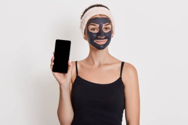 Gülümseyen kız kameraya bakıyor, banyo yaptıktan sonra fotoğraf çekiliyor, siyah t-shirt ve saç bandı takıyor, beyaz arka planda izole edilmiş, boş ekranlı akıllı telefonu gösteriyor..