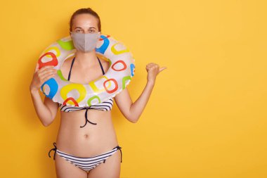 Sarı arka planda izole edilmiş plastik yüzüklü, mayo giyen çekici genç bir kadın, baş parmağıyla işaret ediyor, reklam için fotokopi alanı..
