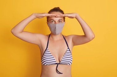 Mayolu ve tıbbi maskeli bir kadın sarı arka planda poz veriyor, avuçlarını alnına yakın tutuyor, mesafeli bakıyor, karantina sırasında koruyucu maske takıyor..