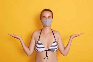 Sarı arka planda şık çizgili mayo ve tıbbi maske takan genç bir kadın iki elini de yana açıp doğrudan kameraya bakıyor..