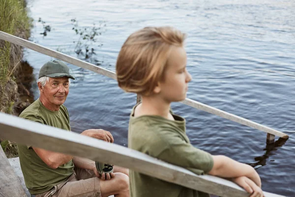 成熟的父亲和他的儿子在户外坐在湖边或河边的木头上 夏天呆在水边 穿着绿色的T恤衫 喜欢一起休息 — 图库照片