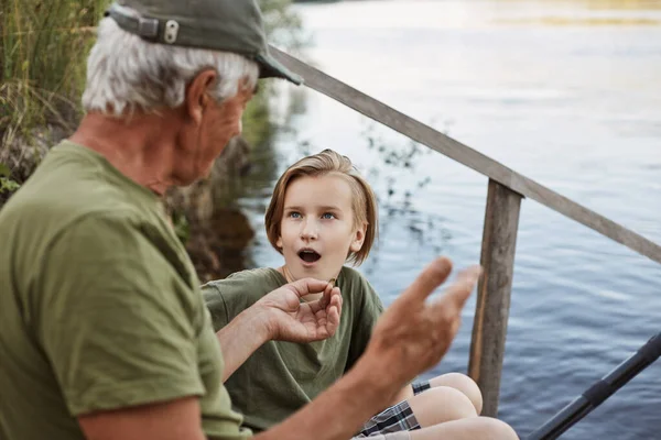 Father and son fishing near river, dad telling about last fishing, showing size of catching fish, son listening with astonished facial expression and opened mouth.