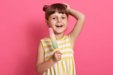 Neşeli çocuk dondurma yiyor ve doğrudan kameraya bakıyor, ellerini başının üstünde tutuyor, mutlu bir şekilde gülüyor, beyaz ve sarı yazlık elbiseler içinde pembe duvarın önünde duruyor..