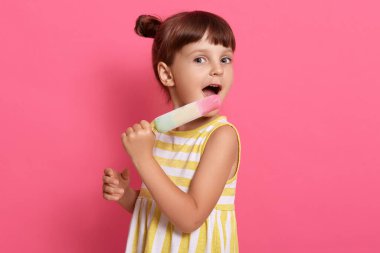 Çocuk pembe arka planda izole bir şekilde poz verirken dondurma yiyor, beyaz ve sarı çizgili yaz elbisesi giyiyor, iki düğüm atıyor, lezzetli şerbetin tadını çıkarıyor..