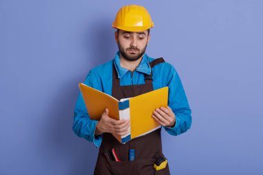 Miğfer ve kahverengi önlük takan, elinde kağıt önlük tutan ve yeni proje hakkında bilgi okuyan konsantre genç mimar ciddi görünüyor, mavi duvara karşı duruyor..
