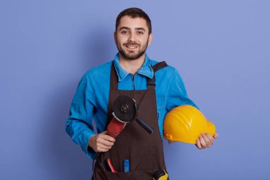 Kahverengi önlük takan, elinde öğütücü alet ve mavi arka planda izole edilmiş sarı koruyucu kaskla gülümseyen sakallı adam kameraya gülümsüyor..