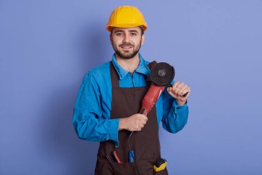Neşeli, yakışıklı, pozitif inşaatçı doğrudan kameraya bakıyor ve içten gülümsüyor, öğütücü ellerde, önlük giyiyor ve koruyucu sarı kask takıyor..