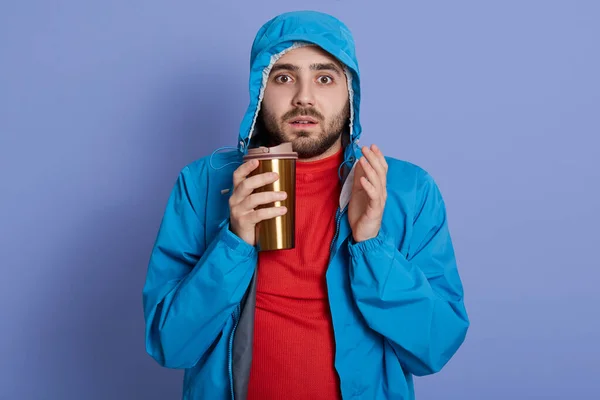 Man wearing jacket and red shirt drinking coffee against blue wall, looking directly at camera with astonished facial expression, being very surprised.