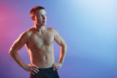 Yakışıklı genç, elleri kalçasında ve doğrudan yan tarafa bakıyor, üstsüz poz veriyor kaslarını gösteriyor, formda duruyor, neon arka planda poz veriyor..