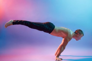 Genç çekici bir adam yoga yapıyor, elleri üzerinde duruyor, üstsüz poz veriyor ve siyah pantolon giyiyor, neon arka planda izole edilmiş, esnek bir adam vücudunu dengeliyor..