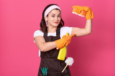 Koyu renk saçlı kadın, pembe arka planda izole olmuş kaslarını gösteriyor. Kahverengi önlük ve beyaz tişört giyen hizmetçi, elinde sünger tutuyor..