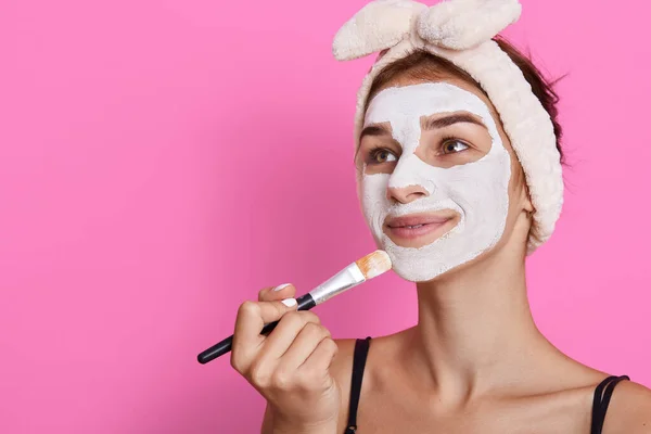 Beautiful young woman applying facial mask at home, looking smiling aside, wearing headband with bow, copy space for advertisement, beauty procedures at home.