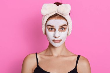 Genç bir kadının yakın plan portresi yüz kilinden maske takıyor, kafasının etrafında beyaz saç bandı var, ciddi görünüyor, pembe arka planda poz veriyor, kameraya bakıyor..