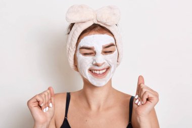 Memnun bir kadının fotoğrafı, gözleri dişlek gülümsemesiyle kapalı, kozmetik prosedürleri var, cilt bakımı için kilden maske takıyor, yaşlanma manipülasyonunu önlüyor..