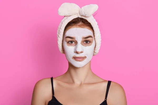 Woman with headband making face moistening procedures, wearing sleeveless t shirt, posing isolated over pink background, looking directly a t camera.