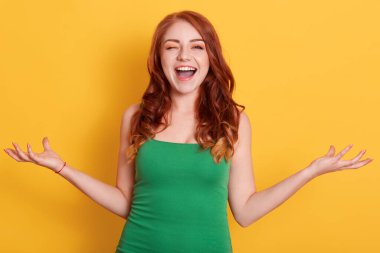 Mutlu bir şekilde poz veren, ellerini yana açıp göz kırpan yeşil elbiseli, sarı arka planda yalnız poz veren bir kadın. Kadın doğrudan kameraya bakıyor..