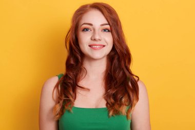 Sarı arka planda gülümseyen genç bir kadın, hoş görünümlü kızıl saçlı, yeşil tişörtlü, kameraya bakıyor, pozitif duygularını ifade ediyor..