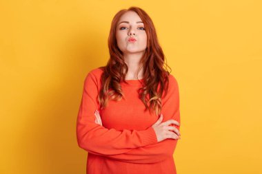 Kendine güvenen kızıl saçlı, ağzı açık turuncu kazak giyen öpüşme hareketi gösteren dudaklarını yuvarlak tutan, sarı duvarın üzerinde tek başına duran bir kadın..