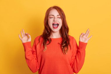 Neşeli yüzlü genç bir kadın, sarı arka planda yalnız poz veriyor, gözlerini kapalı tutuyor ve normal turuncu süveter giyiyor, iki eliyle ok işaretleri gösteriyor..