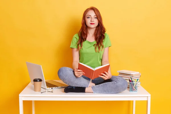 红头发的女学生两腿交叉坐在桌子上 手里拿着书本 看着相机 对远距离学习感到厌烦 穿着休闲装 — 图库照片