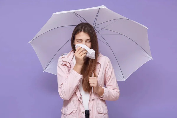 Young woman in rose raincoat covering mouth with handkerchief, posing under white umbrella isolated over lilac wall background, having runny nose, being sick, wet weather.