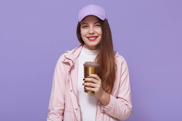 Pretty dark haired European woman has long hair, wears jacket and baseball cap, holds coffee to go in thermo mug, models over lilac wall, looks smiling at camera.