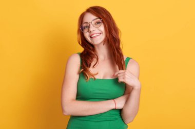 Mutlu gülümseyen kızıl saçlı güzel kadın yeşil kolsuz tişört giyiyor, büyüleyici bir gülümsemeyle kameraya bakıyor, sarı arka planda tek başına duruyor..