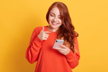 Neşeli kadın işaret parmağıyla akıllı telefon ekranını işaret ediyor, cep telefonuna büyüleyici bir gülümsemeyle bakıyor, sarı arka planda tek başına duruyor..