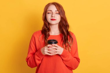 Sakin bir kadın, sıradan bir süveter giyiyor, iki elinde hazır kahve tutuyor, sıcak içkinin tadını çıkarıyor, gözleri kapalı, sarı arka planda başarılı bir kız..