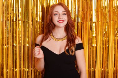 Genç, enfes, zarif, kızıl saçlı, siyah elbise giyen, altın sarısı süslemelerle süslenmiş, kırmızı dudaklı mutlu kadın, siyah elbise giyiyor, partide..