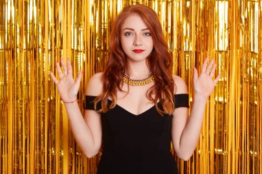 Girl being pissed and losing temper while posing against wall with golden tinsel, european red haired girl with beautiful curls, looks angry, raised hands up, being offended during party.