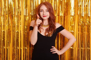 Gülümseyen genç bir kadın işaret parmağıyla kameraya işaret ediyor ve gülümsüyor, bayramı kutluyor, siyah elbiseler içinde, duvarın üzerinde izole edilmiş, altın süslemelerle süslenmiş, partide güzel kızıl saçlı bir kız..