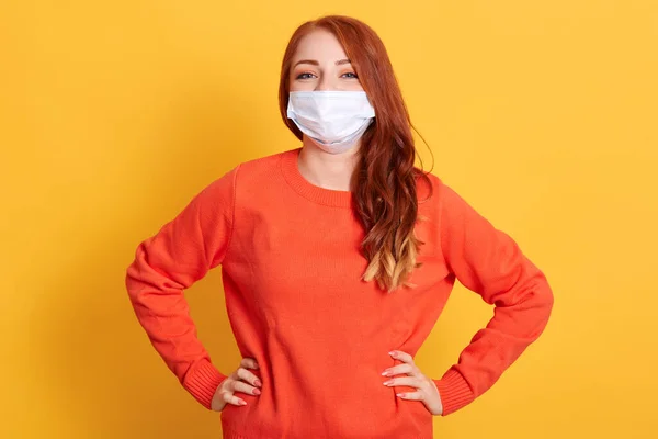 여자는 의료용 마스크와 주황색의 캐주얼 스웨터를 카메라를 쳐다보며 노란색 배경에 — 스톡 사진