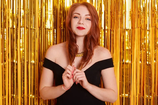 Kızıl saçlı, kibar ve güzel, arka planda altın sarısı süslemeli bir kız, kameraya bakıyor, buklelerine dokunuyor, çıplak omuzlu zarif bir elbise giyiyor..
