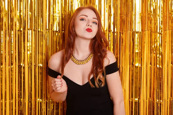 Kırmızı dudaklarıyla poz veren, öpüşme jesti yapan, altın sarısı duvara poz veren, siyah elbise giyen, siyah kolyeli, zarif Avrupalı kızıl saçlı bir kadın...