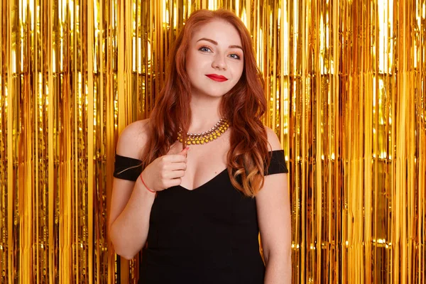 Siyah elbise ve kolye giyen sevimli kızıl saçlı kız, arka planda altın sarısına karşı duruyor, sakin bir ifadeyle kameraya bakıyor, partiyi kutluyor..