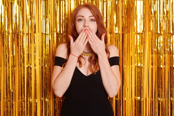 Beautiful red haired happy woman at celebration birthday party, New Year eve celebrating, being surprised, cover mouth with palms, looks at camera, wearing elegant black dress.