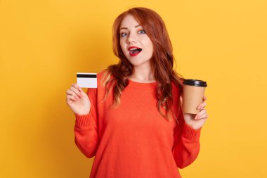 Astonished buyer finding offer online, holding take away coffee and credit card, has surprised facial expression, lady with red lips and wavy hair, wearing orange sweater. clipart