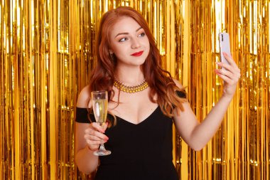 Siyah elbiseli ve kolyeli kızıl saçlı kadın akıllı telefonuyla selfie çekiyor, elinde şarap kadehiyle duvarın üzerine izole edilmiş bir şekilde poz veriyor..