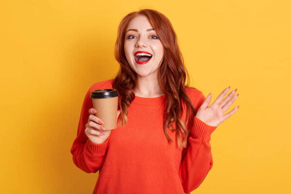 Pretty red haired female prefers coffee take away, holds disposable cup with aromatic hot drink, looking at camera with glad expression and toothy smile, wears orange sweater.