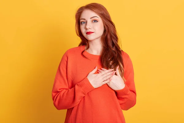 Lovely female with friendly look, keep hands on chest, expresses positive and good attitude, has red wavy hair, feels gratitude to her mother, posing isolated over yellow background.