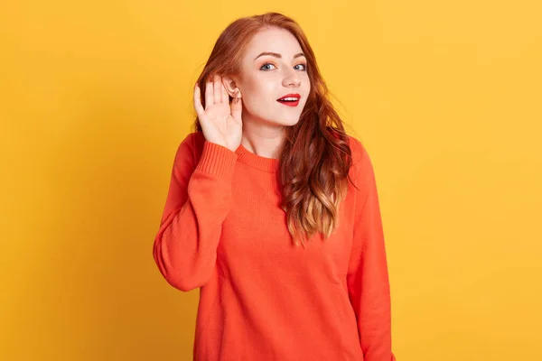 Funny astonished red haired European female, raising eyebrows, expressing surprise, keeping hand near ear trying to listen to gossips, wearing orange casual sweater.