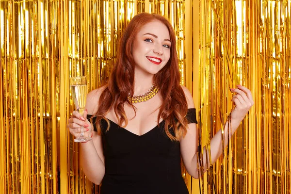 Altın süslemelerle süslenmiş duvara poz veren, siyah elbise ve kolye takan kızıl saçlı, bayramı kutlayan gülümseyen bir kadın portresi..